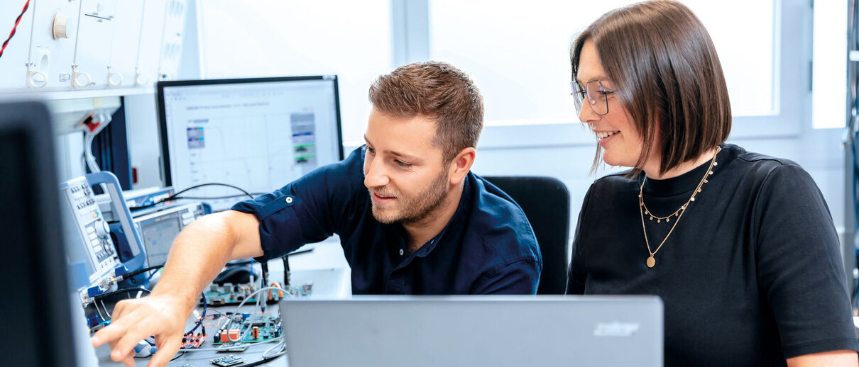 Mitarbeiter und Mitarbeiterin sitzen an einem Arbeitsplatz mit elektronischen Messgeräten und Computern