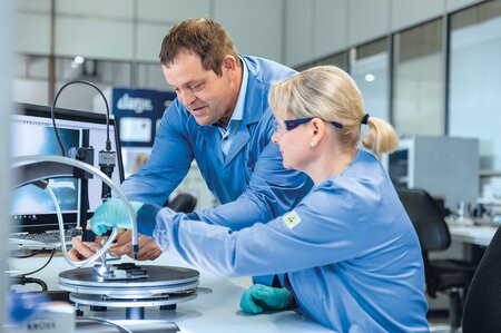 Zwei Produktionsmitarbeiter Mitarbeiter der Zollner Elektronik AG führen eine Kontaktwinkelmessung an einem Prüfgerät zur Bestimmung der Benetzbarkeit von Feststoffoberflächen durch.