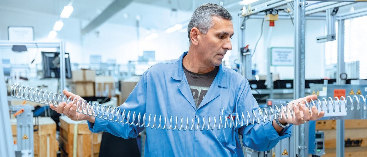 Mitarbeiter der Zollner Elektronik AG hält große Metallfeder in moderner Produktionshalle.