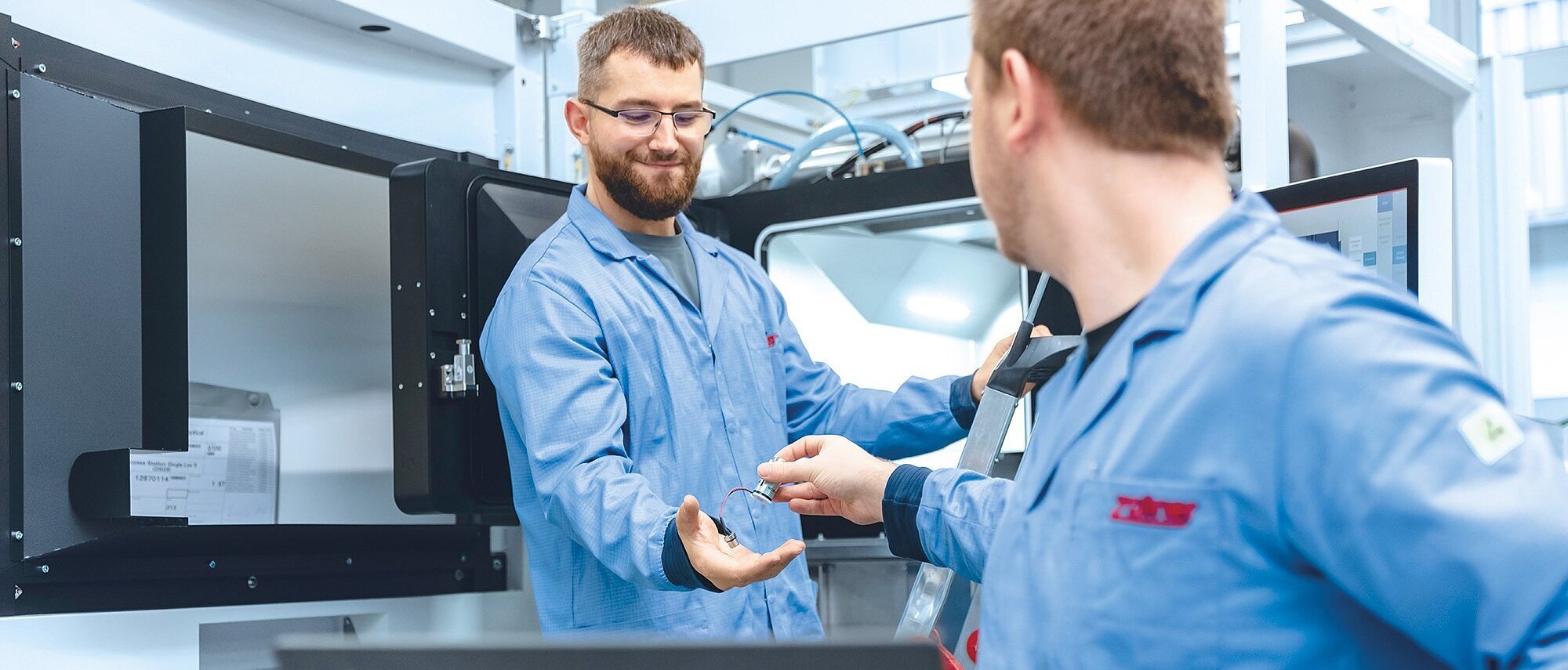 Zwei Produktionsmitarbeiter stehen bei einer Maschine, der eine überreicht dem anderen etwas 