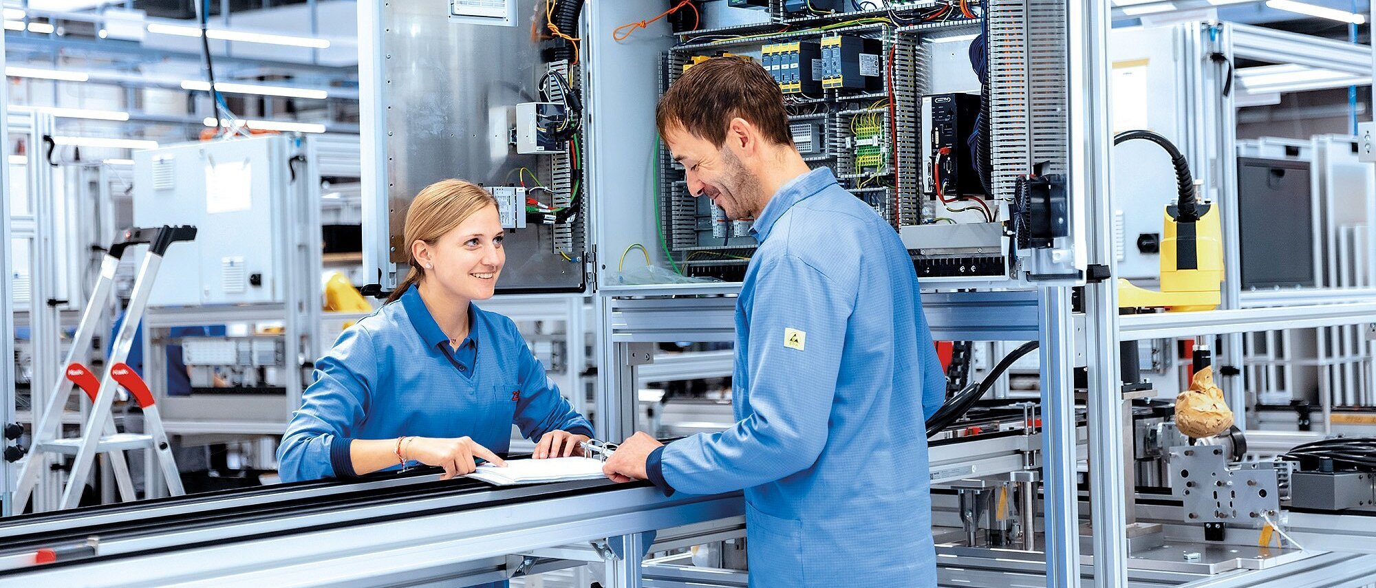 Zwei Mitarbeitende in ESD-Schutzkleidung stehen vor einer Fertigungslinie und besprechen Arbeitsunterlagen in einer modernen Produktionshalle