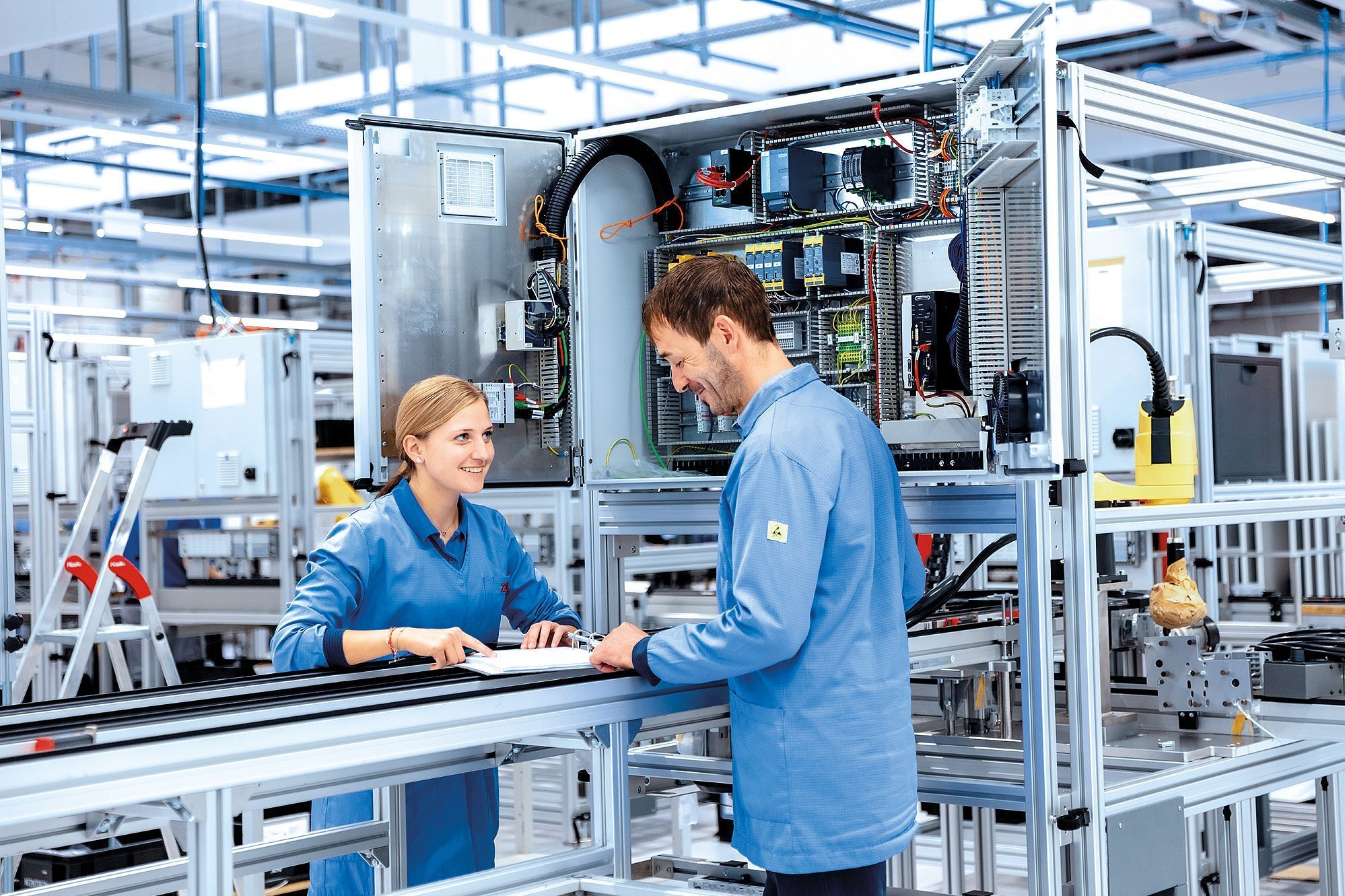 Zwei Mitarbeitende in ESD-Schutzkleidung stehen vor einer Fertigungslinie und besprechen Arbeitsunterlagen in einer modernen Produktionshalle