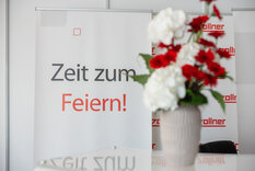 Bild von einem weiß/roten Blumenstrauß und im Hintergrund ein RollUp mit der Aufschrift "Zeit zum Feiern" 