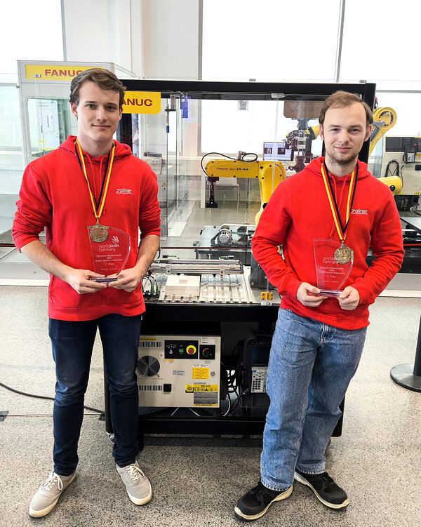 Zwei Mitarbeiter der Zollner Elektronik AG präsentieren Siegerpokale und Medaillen vor einer FANUC-Roboteranlage.