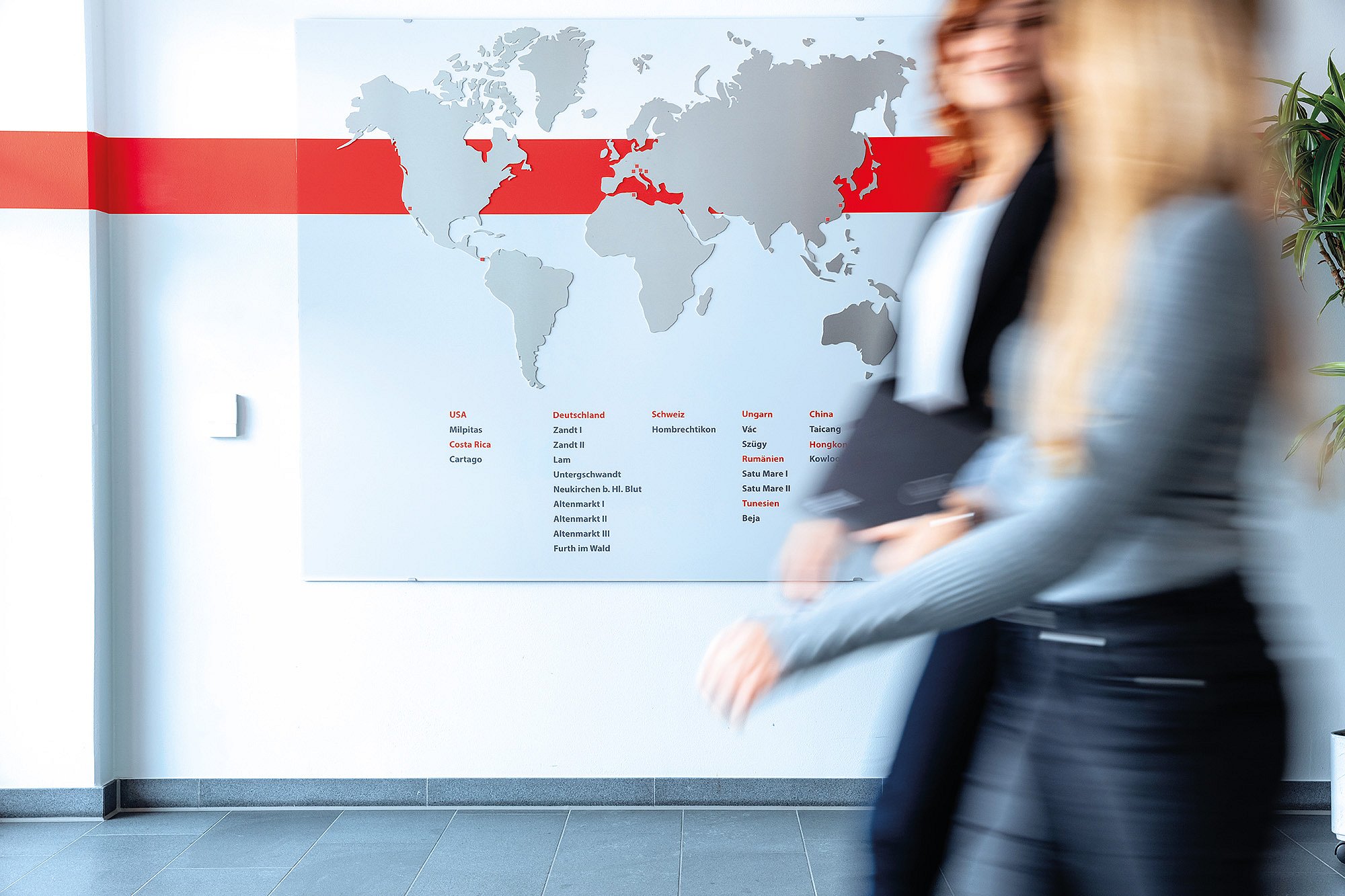 Zwei Mitarbeiterinnen der Zollner Elektronik AG gehen durch einen Flur vor einer Weltkarte im Bereich Global Procurement.